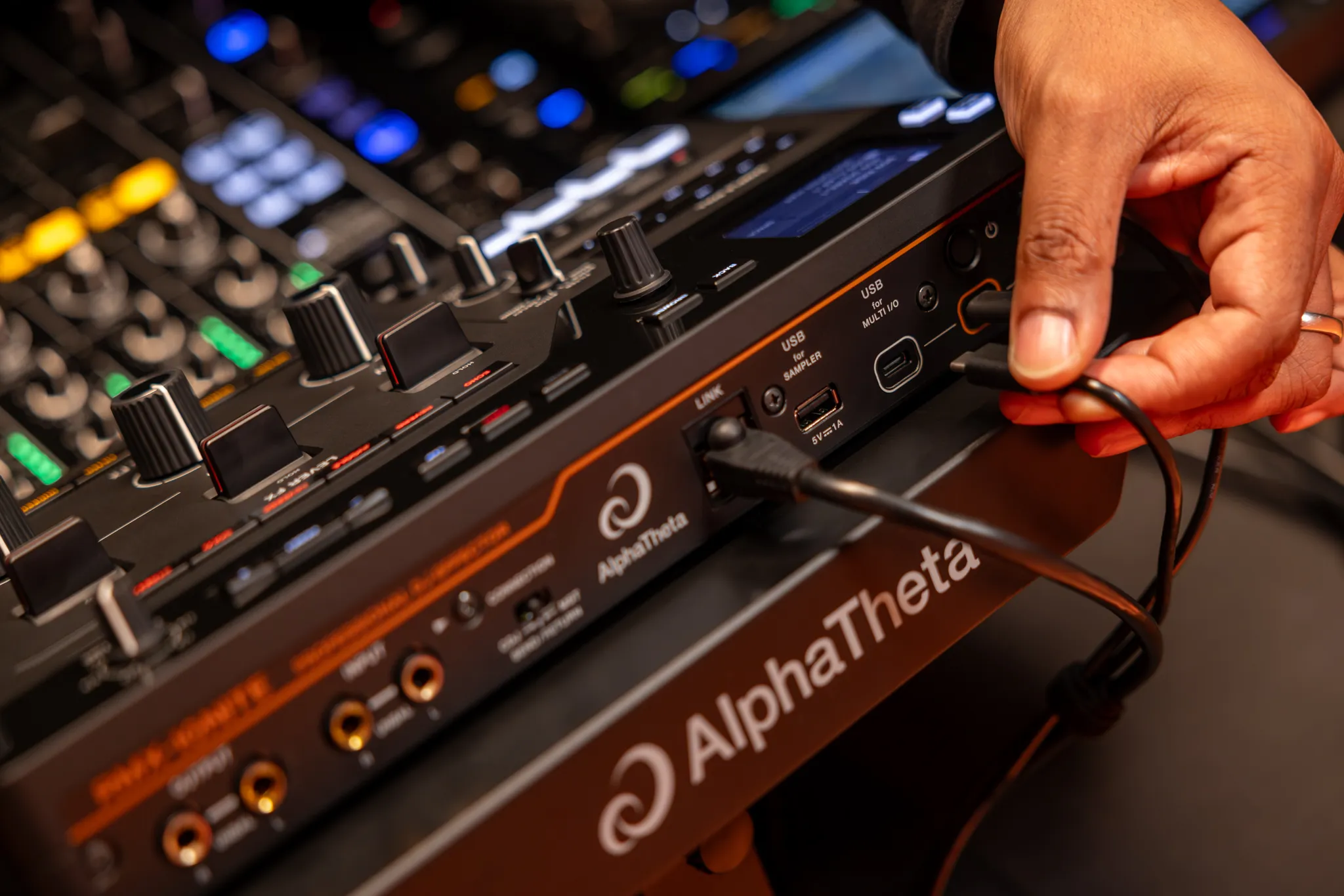 Detalle de las manos de un DJ conectando cables USB y de alimentación en el panel posterior de la unidad de efectos AlphaTheta RMX-IGNITE.